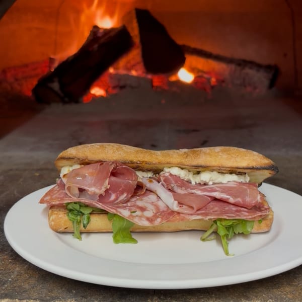 A Maeposas Italian sandwich with cured meats and greens on a white plate, in front of a wood-fired oven.