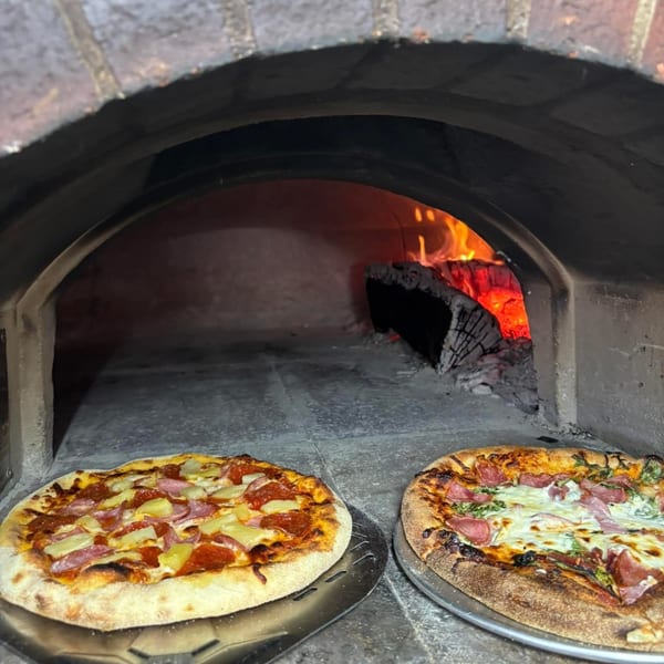 Two Maeposas pizzas, one with pineapple and pepperoni, sit before a wood-fired oven.