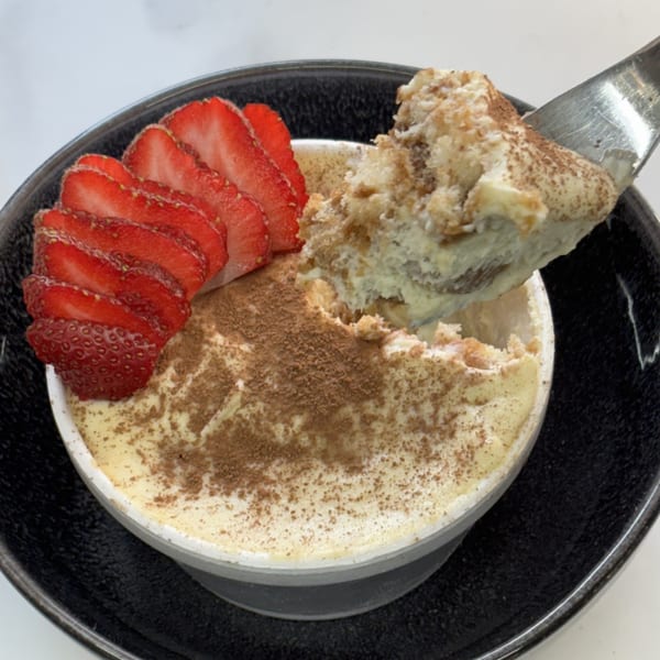 Tiramisu dessert with cocoa powder and sliced strawberries in a black bowl, with a spoon taking a scoop.